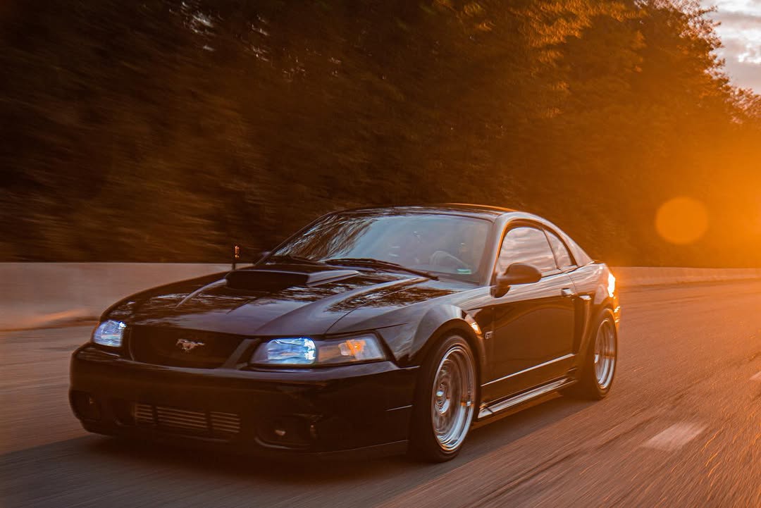 A black Mustang on the road at golden hour with its headlights on